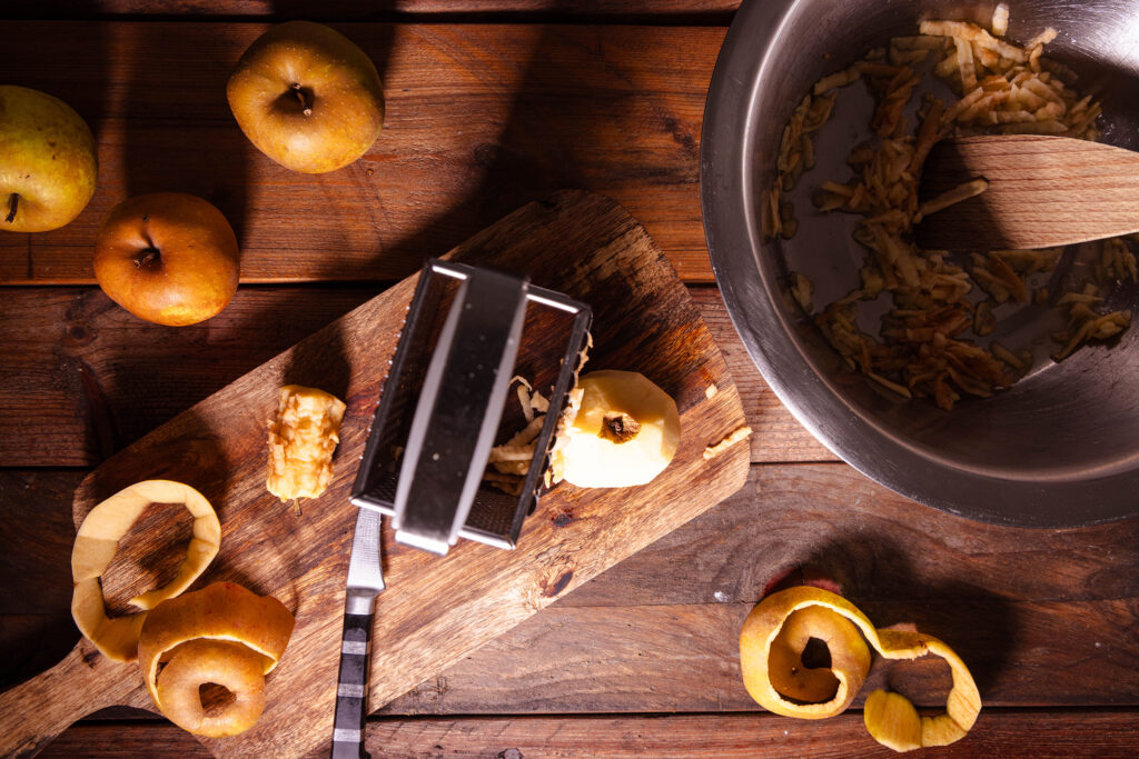 Äpfel, Apfelschalen, eine Vierkantreibe und eine Schüssel mit geriebenen Äpfeln.