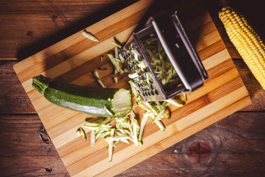 Eine Zucchini wird mit einer Vierkantreibe auf einem Holzbrett gerieben.