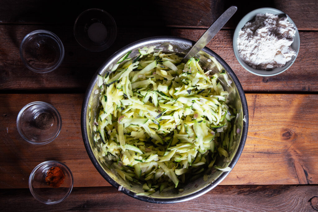 Eine Schüssel mit geriebenen Zucchini, Kartoffeln, gewürfelten Schallotten und angerührtem Kichererbsenmehl.