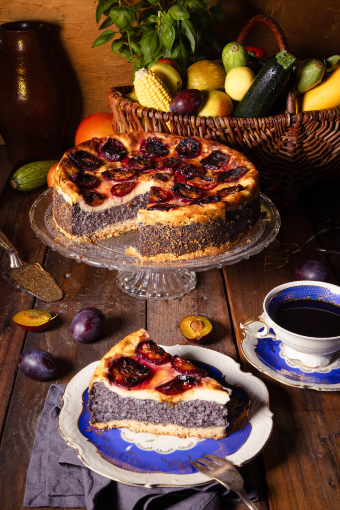 Zwetschen-Mohn-Käsekuchen auf einem alten Holztisch mit einem Korb voller saisonaler Früchte und Gemüse.