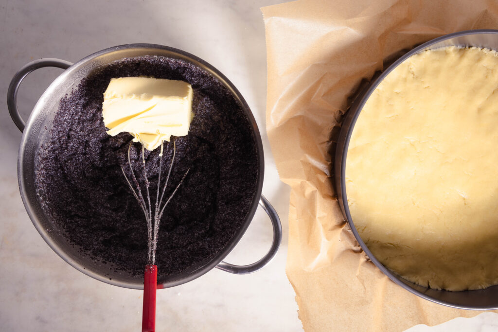Eine Springform mit Mürbeteig und ein Topf mit Mohnfüllung. Auf der Mohnfüllung ein Stück vegane Butter.