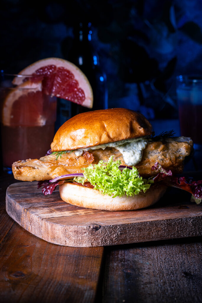 Veganer Fischburger auf einem Holzbrett, dahinter ein Getränk