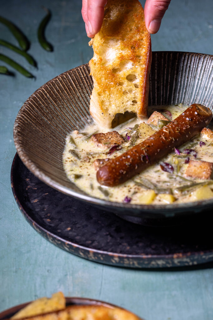 Ein Stück geröstetes Brot wird in eine Schale Schnippelbohnensuppe getunkt.