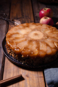 Überkopf-Apfelkuchen auf einem Kuchenteller, dahinter frische, rote Äpfel