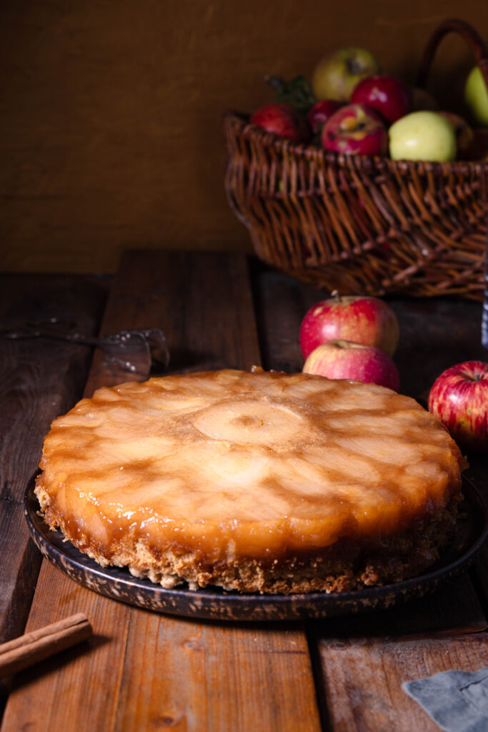 Überkopf-Apfelkuchen auf einem Kuchenteller, dahinter frische, rote Äpfel