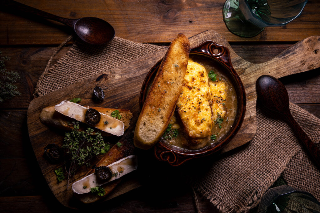 Zwiebelsuppe, französische Art – hier mit gerösteten Baguettescheiben als Beilage. Teilweise belegt mit meinem Blumenbert, selbstgemachtem Camembert aus Blumenkohl, und süß-sauer eingelegten grünen Walnüssen.