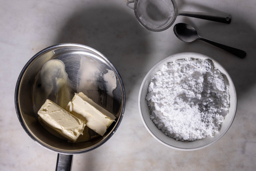 Vegane Butter in einem kleinen Kochtopf, daneben eine Schale Puderzucker.