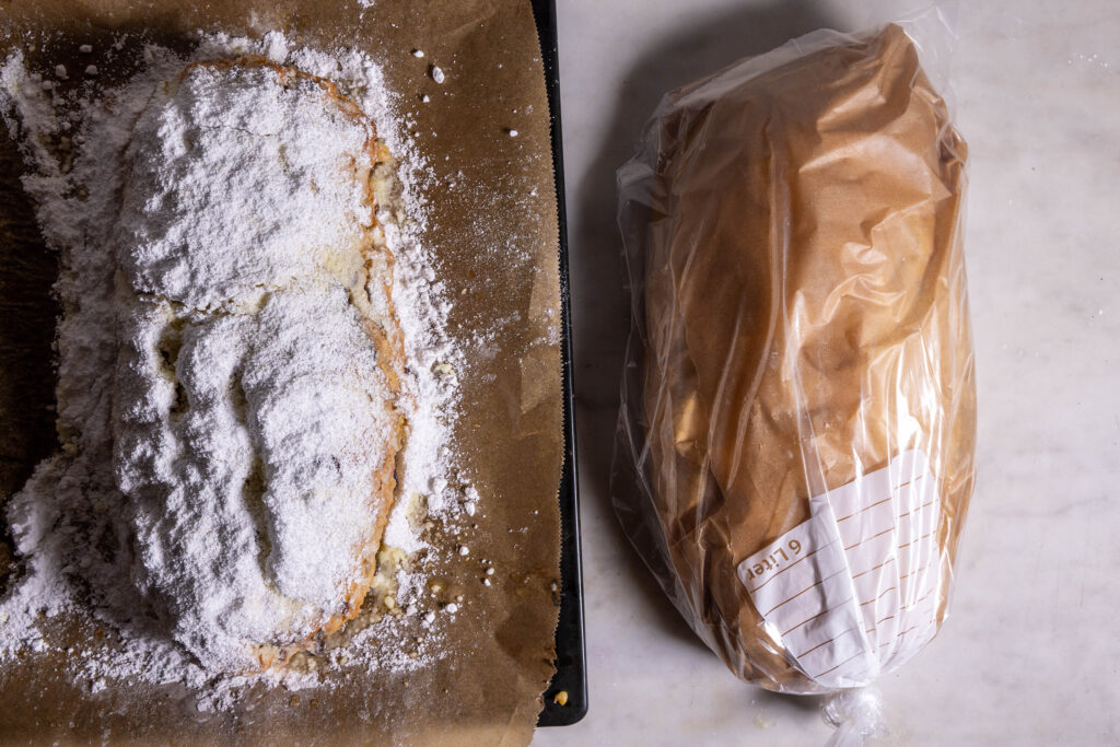Ein veganer Christstollen auf einem Blech, dick mit Puderzucker bestäubt. Ein Stollen in Backpapier und Gefrierbeutel eingepackt daneben.