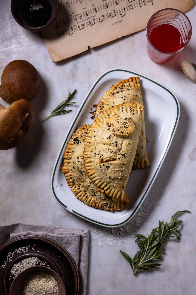 Hand Pies mit Waldpilzfüllung – drei auf einem Teller, darum zwei Steinpilze, ein Notenblatt, ein Getrränk.