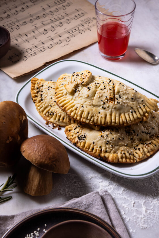 Hand Pies mit Waldpilzfüllung – drei auf einem Teller, darum zwei Steinpilze, ein Notenblatt, ein Getrränk.