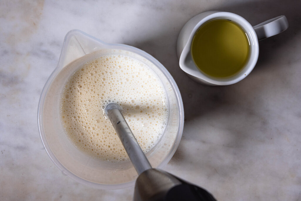 Vegane Mayonnaise aus Sojamilch wird in einem Messbecher püriert. Daneben eine Karaffe Öl.