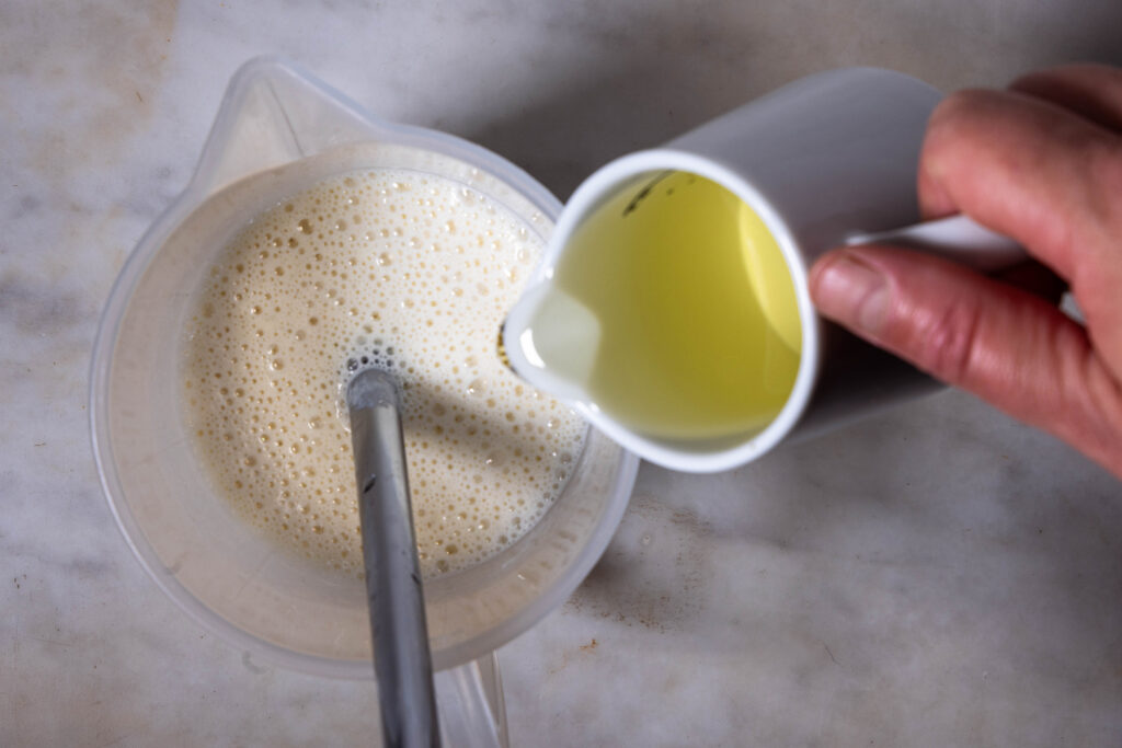 Vegane Mayonnaise aus Sojamilch wird in einem Messbecher püriert. Aus einer Karaffe wird etwas Öl hinein gegossen.