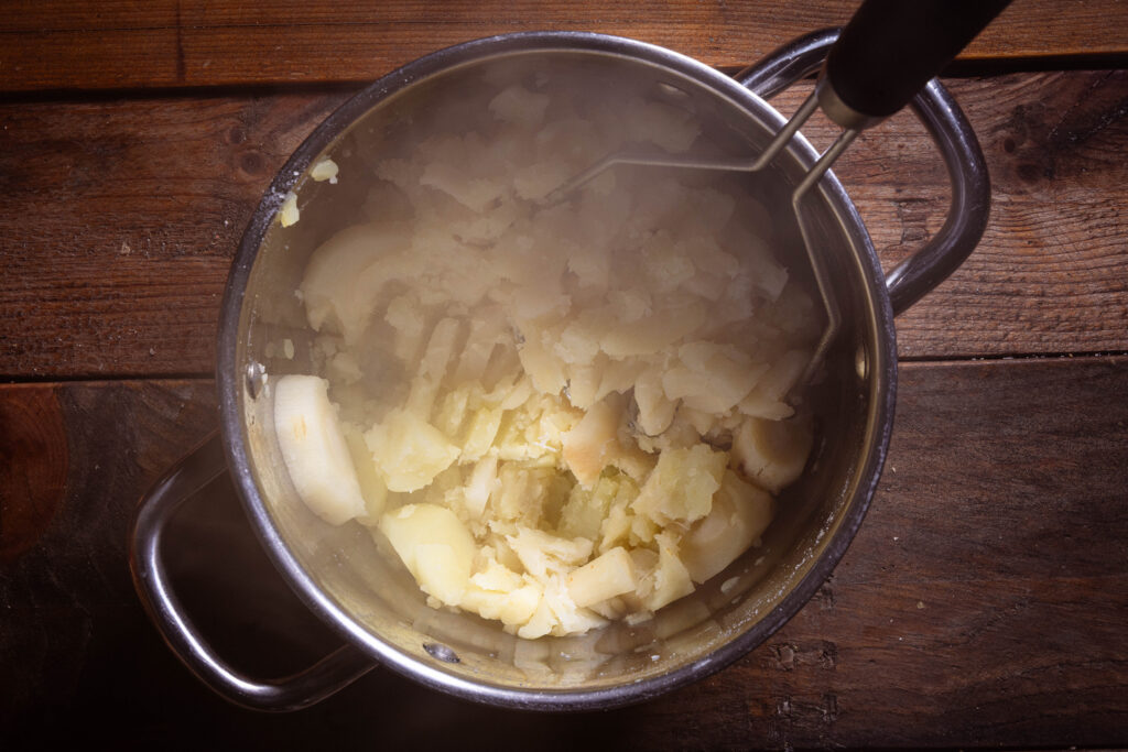 Ein Kartoffelstampfer stampft dampfende Pastinaken und Kartoffeln zu Püree.