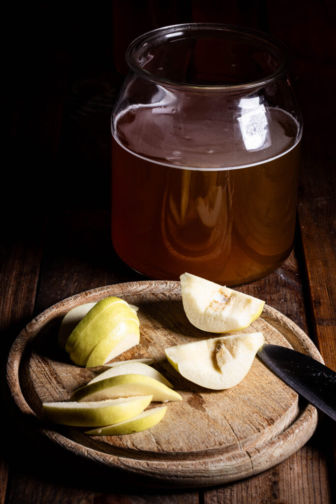 Quittenviertel und Quittenschnitze auf einem Holzbrett. Dahinter ein Gefäß mit Kombucha.