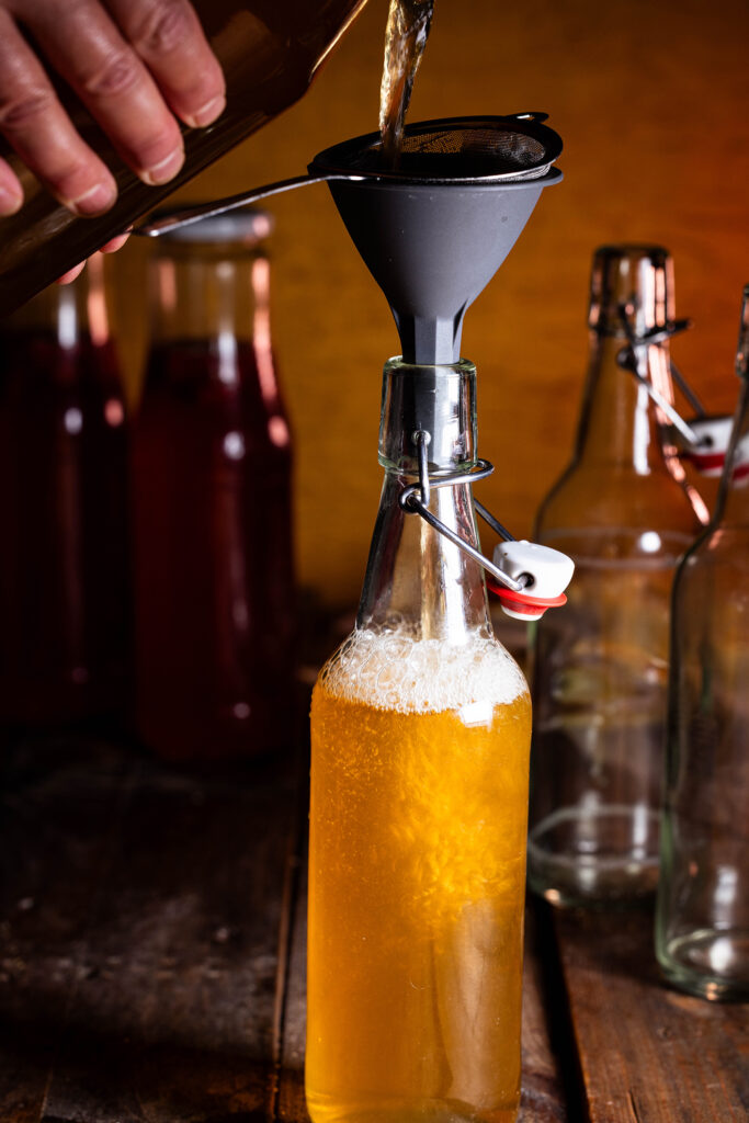 Quitten-Kombucha wird mit einem Trichter und einem Sieb in eine Flasche abgefüllt.