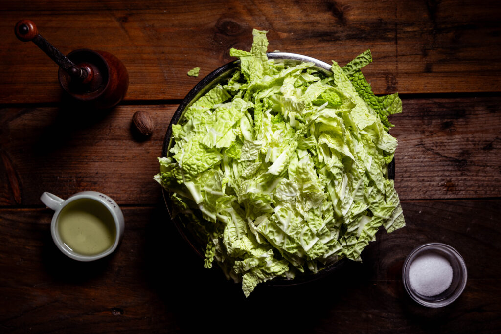 Geschnittener Wirsing in einer Schüssel, außerdem Salz, Pfeffer, eine Muskatnuss und Öl auf einem Holztisch.