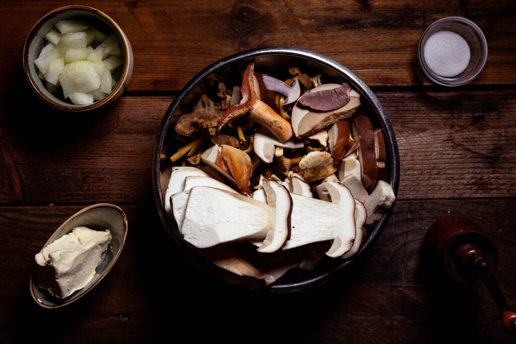 Eine Schale mit geputzten Waldpilzen, in Scheiben geschnitten. Außerdem Zwiebelwürfel, Salz, Pfeffer und vegane Butter.
