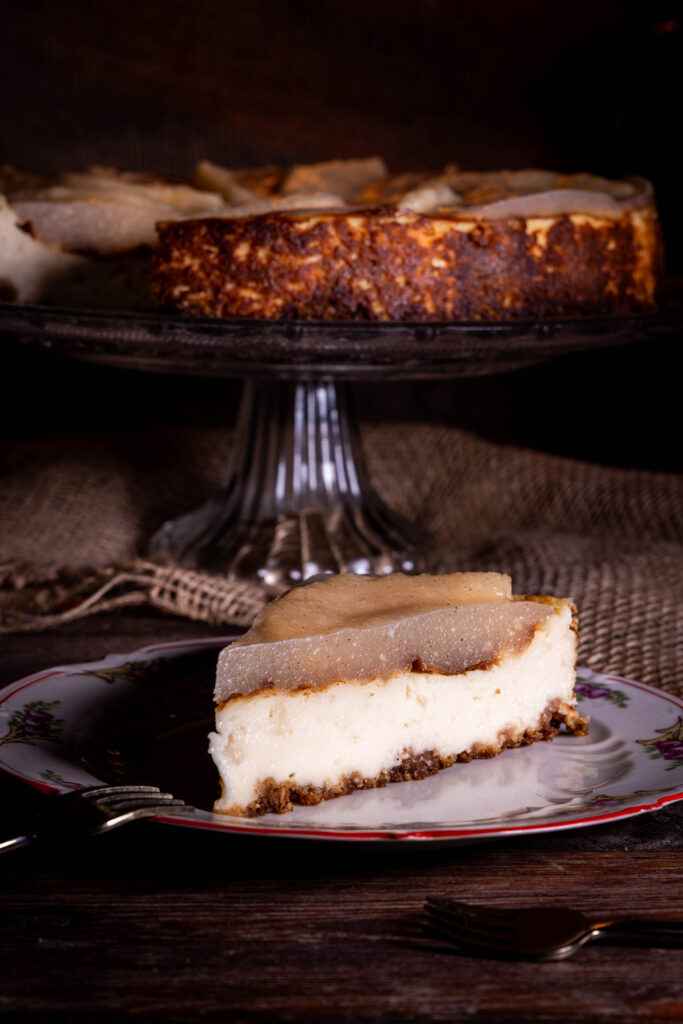 Veganer Käsekuchen mit fermentierten Quitten, ein Stück auf einem Teller, der Rest des Kuchens dahinter auf einer Etagere