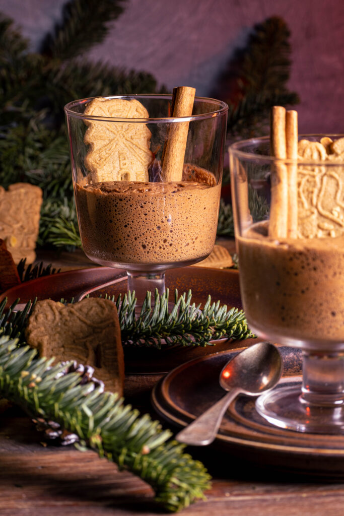 Spekulatius-Mousse in Dessertgläsern, dekoriert mit Zimtstange und einem Spekulatiuskeks. Auf einem weihnachtlichen Holztisch.