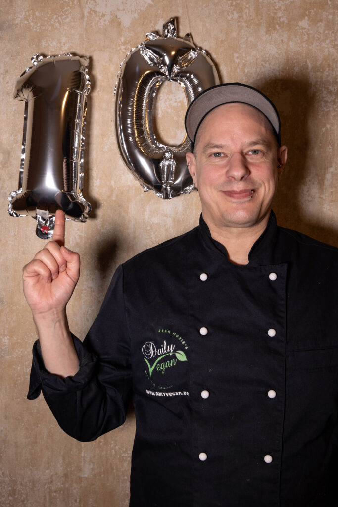 Sean Moxie feiert 10 Jahre Daily Vegan. Hinter ihm eine 10 aus silbernen Luftballons, auf die er zeigt.