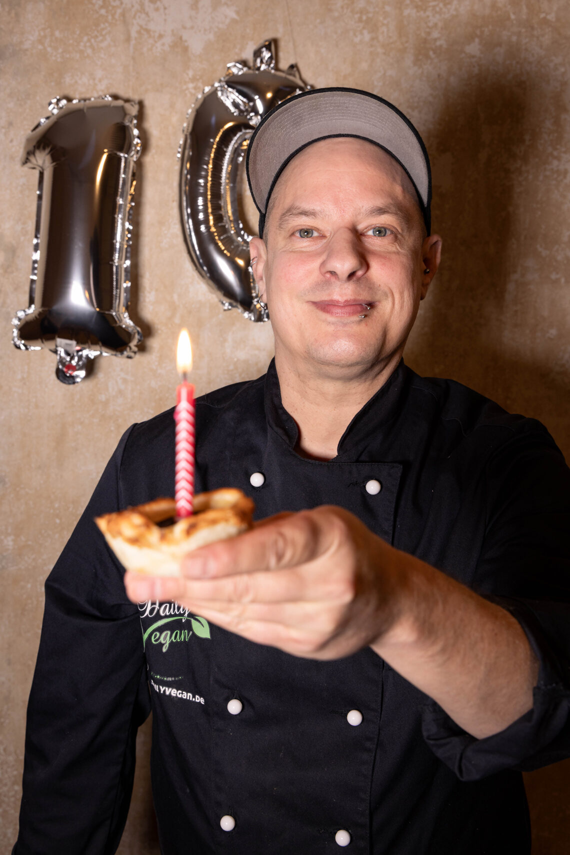 10 Jahre Daily Vegan. Sean Moxie mit einer Pasteis de Nata mit Kerze darin, HInter Ihm eine 10 aus silbernen Luftballons.