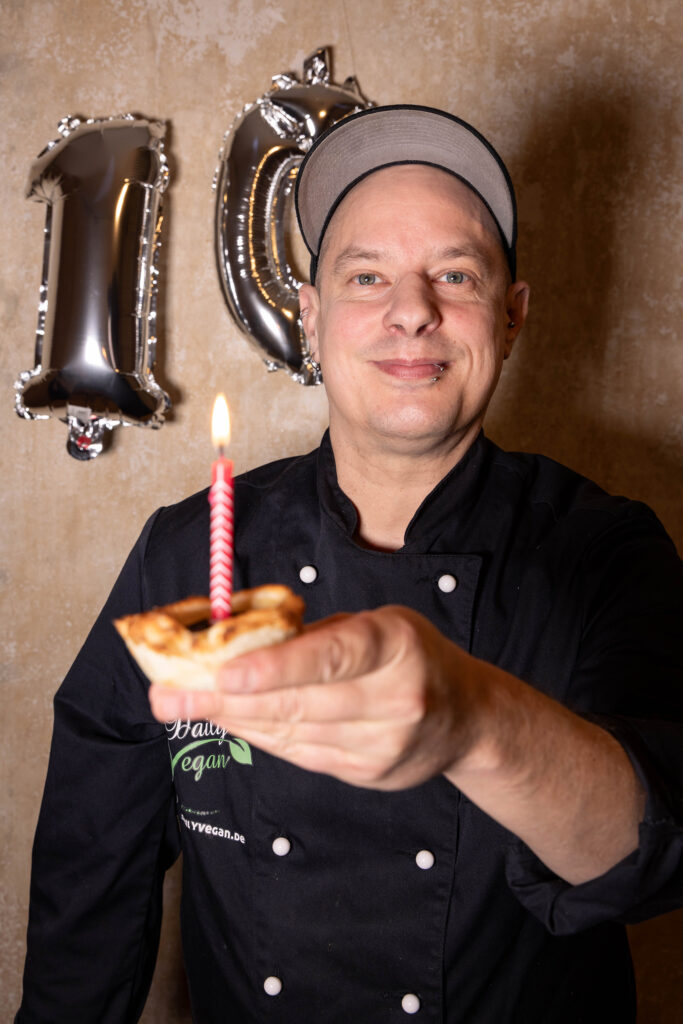 10 Jahre Daily Vegan. Sean Moxie mit einer Pasteis de Nata mit Kerze darin, HInter Ihm eine 10 aus silbernen Luftballons.