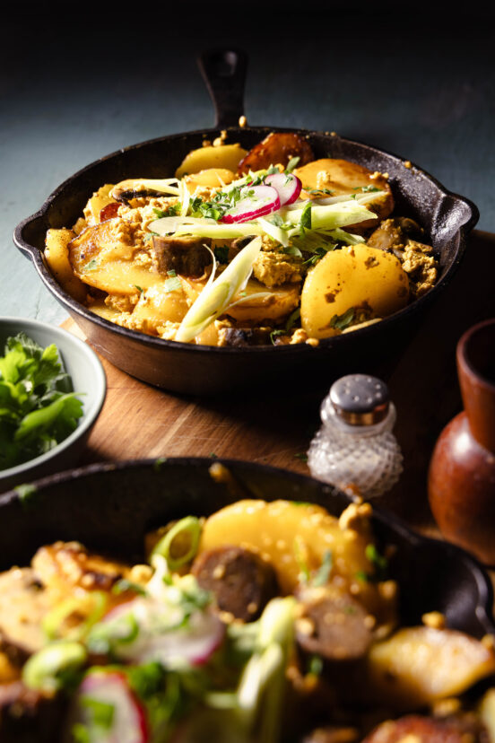 Veganes Bauernfrühstück mit Würstchen und Pilzen in zwei gusseisernen Pfannen auf einem Holzbrett.
