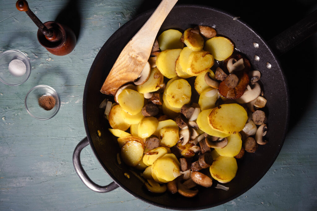 Bratkartoffeln, vegane Würstchen, Champignons und Zwiebeln braten in einer Pfanne.