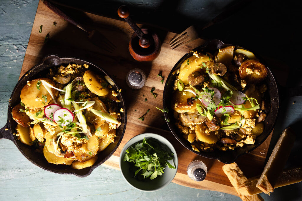 Veganes Bauernfrühstück mit Würstchen und Pilzen in zwei gusseisernen Pfannen auf einem Holzbrett.