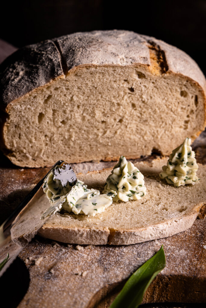 Vegane Bärlauchbutter, zu Rosetten gespritzt auf selsbtgebackenem Brot. Eine Butterrosette wird mit einem Messer verstrichen.