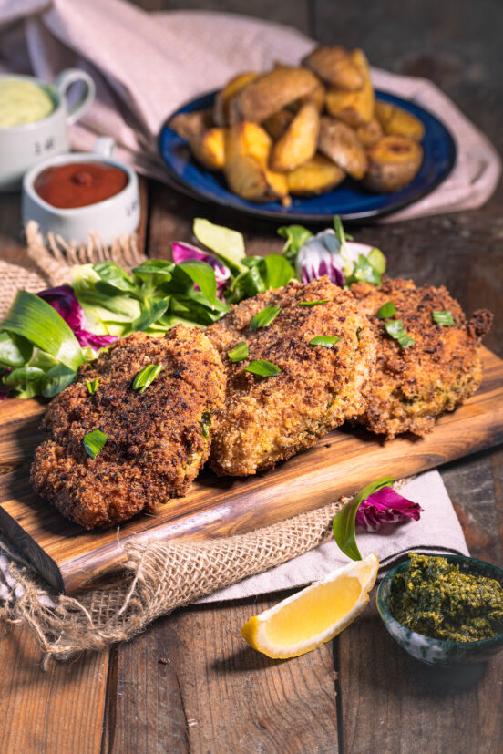 Mit Bärlauch gefülltes Schnitzel in Röstzwiebelpanade - vegan - 3 Stück auf einem Holzbrett. Dahinter Wedges auf einem Teller.