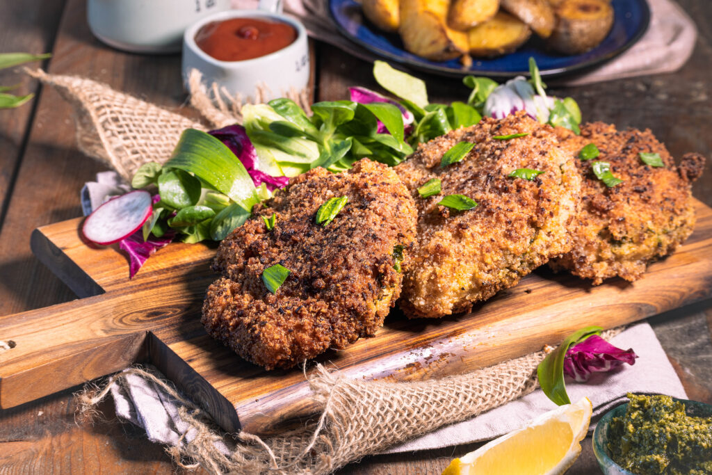Mit Bärlauch gefülltes Schnitzel in Röstzwiebelpanade - vegan - 3 Stück auf einem Holzbrett. Dahinter Wedges auf einem Teller.