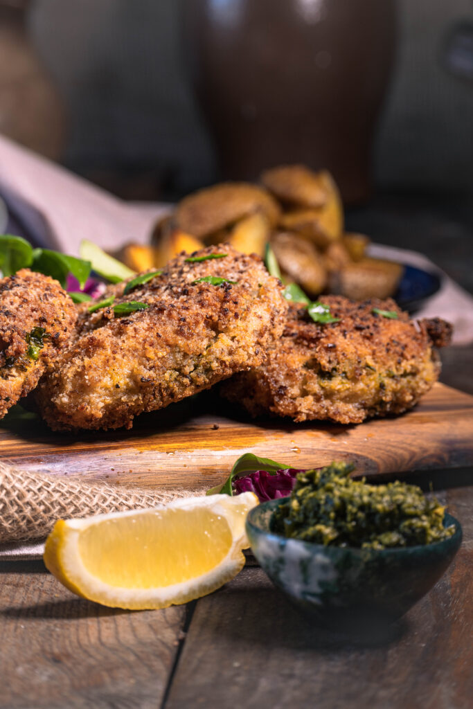 Mit Bärlauch gefülltes Schnitzel in Röstzwiebelpanade - vegan - 3 Stück auf einem Holzbrett. Dahinter Wedges auf einem Teller.