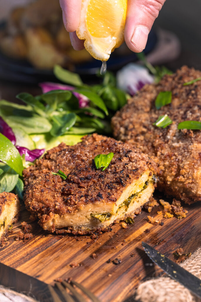 Mit Bärlauch gefülltes Schnitzel in krosser Zwiebelpanade - angeschnitten auf einem Holzbrett. Darüber wird Zitronensaft aus einem Zitronenschnitz gegeben.