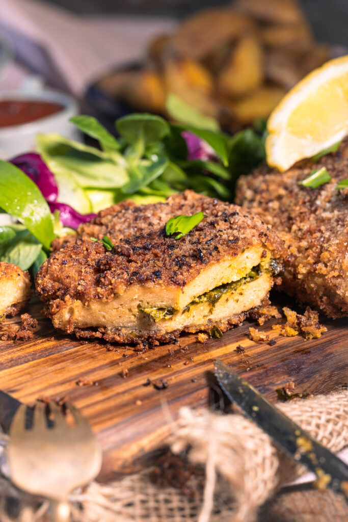 Mit Bärlauch gefülltes Schnitzel in krosser Zwiebelpanade - angeschnitten auf einem Holzbrett. Dahinter Salat.