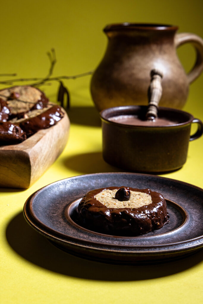 Haselkätzchenplätzchen auf einem Teller, dahinter eine Tasse Kakao, mehr Plätzchen und eine Karaffe.