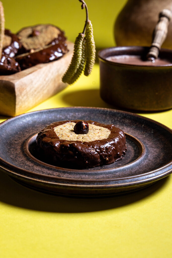Haselkätzchenplätzchen auf einem Teller, dahinter eine Tasse Kakao, mehr Plätzchen und eine Karaffe.