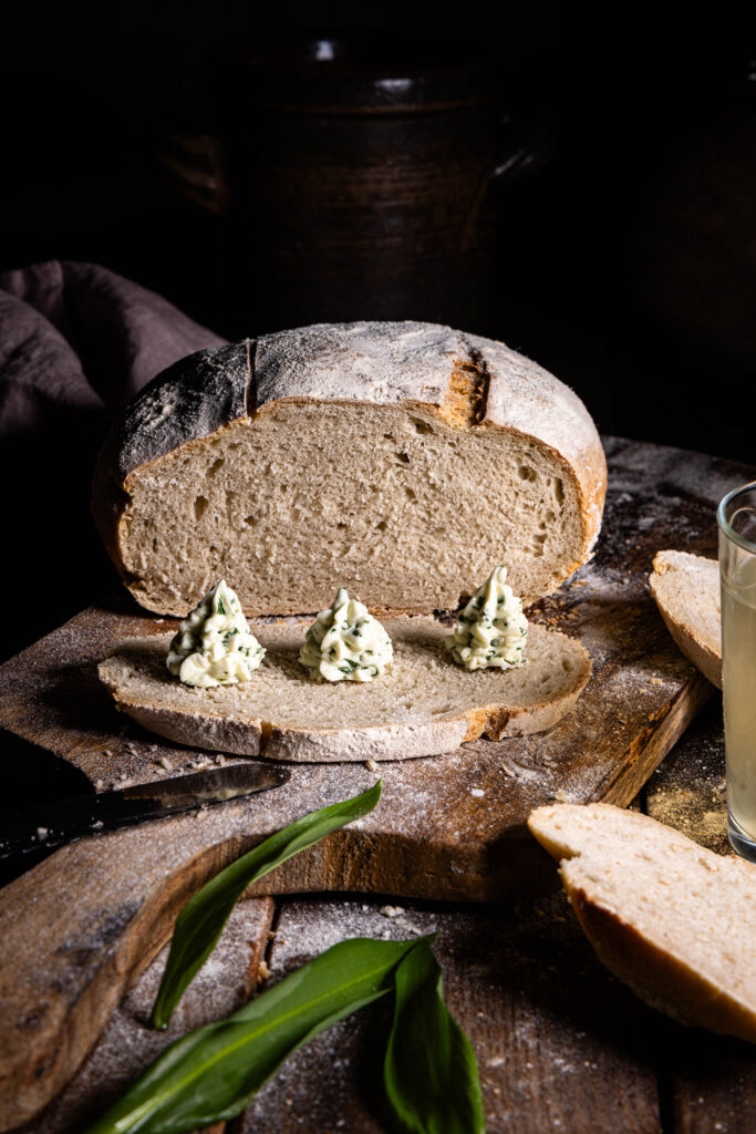 Ganz puristisch genießen: Sojakefir-Roggenbrot mit Bärlauchbutter. Ein angeschnittenes Brot, davor eine Scheibe mit drei gespritzten Rosetten Bärlauchbutter darauf.