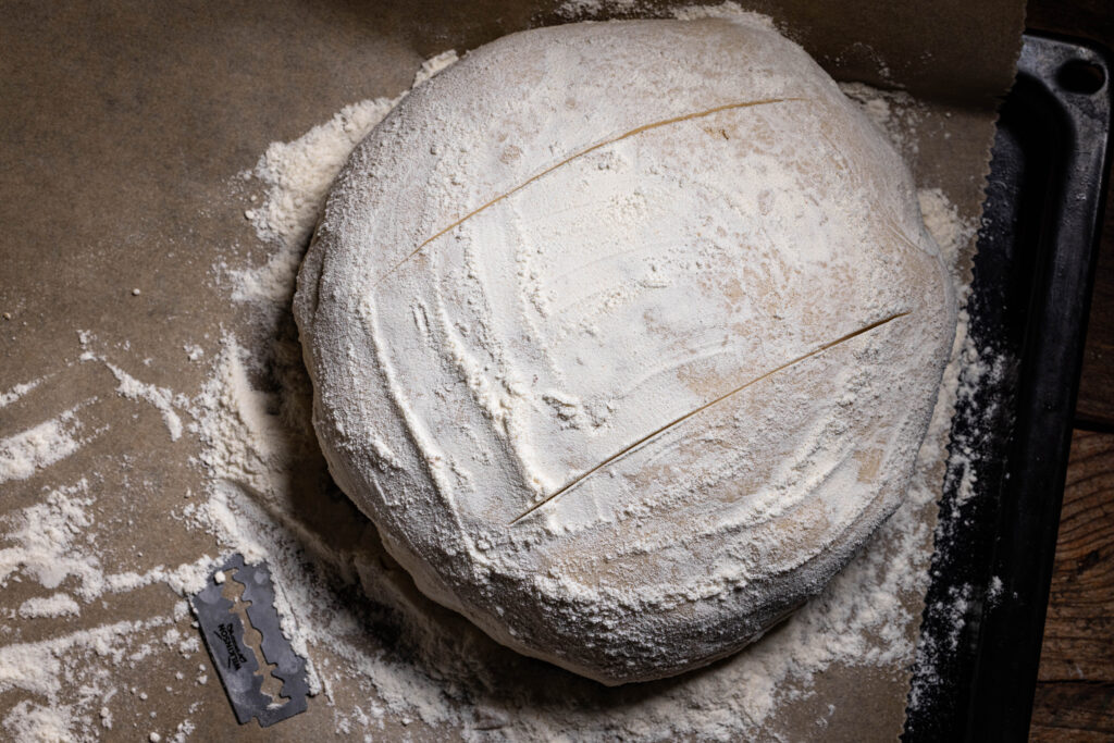 Ein Kefir-Roggenbrot liegt ungebacken auf einem Backblech, außerdem liegt darauf eine Rasierklinge vom Einritzen des Brots.