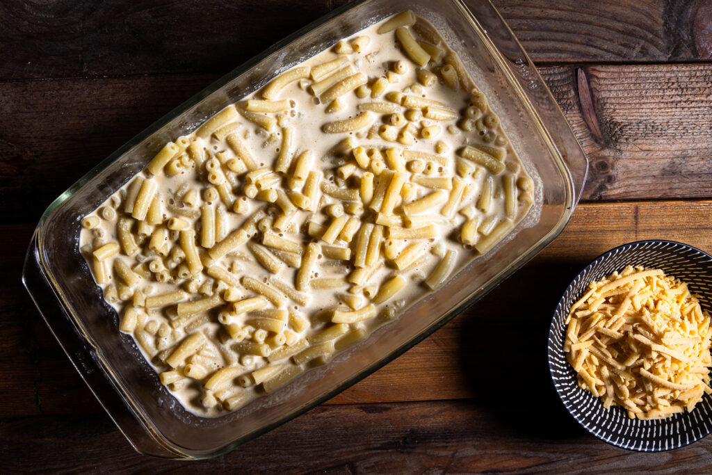 Makkaroni und vegane Käsesauce in einer Auflaufform vermengt. Daneben eine Schale mit veganem, geriebenen Käse.