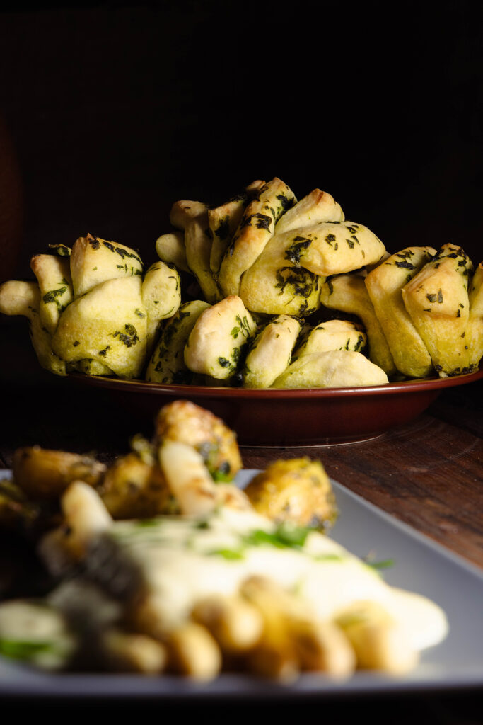Ein Teller mit Bärlauchzupfbroten. Davor verschwommen zu sehen: ein Teller mit Spargel, Kartoffeln und Sauce Hollandaise.