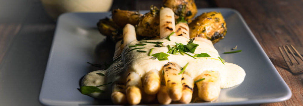 Gegrillter, weißer Spargel mit veganer Sauce Hollandaise, Kräutern und Ofenkartoffeln auf einem Teller.