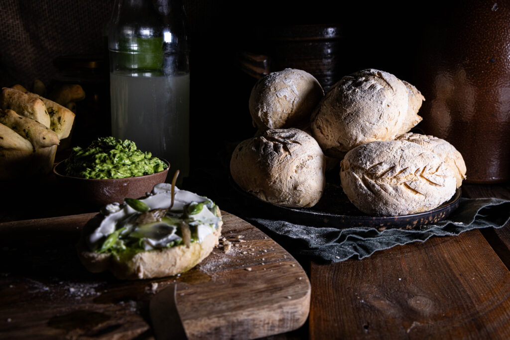 Kefir-Dinkel-Brötchen auf einem Teller, eine Hälfte bestirchen uaf einem Holzbrett. IM HIntergrund Wasserkefir.