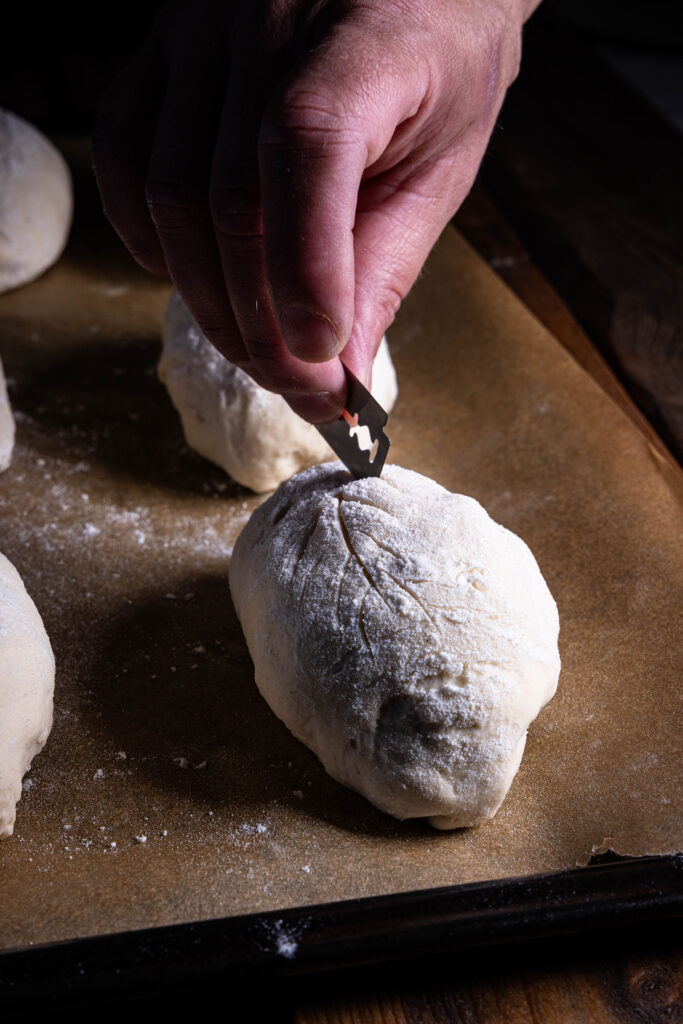 Ein rohes Dinkel-Kefir-Brötchen wird mit einer Rasierklinge eingeritzt.