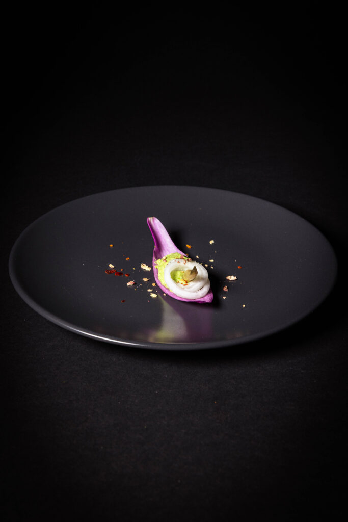 Amuse Bouche für den Frühling - Magnolienblüte, Erbse, Mairübchen, Bärlauchknospe - auf einem schwarzen Teller.